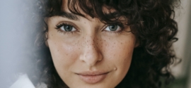 Headshot of attractive female with dark curly hair looking at camera against green leaves on blurred background in light room
