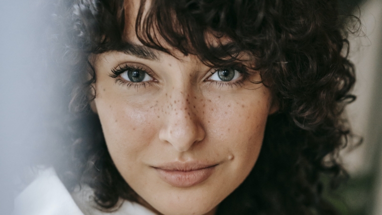 7 Ways to Look More Feminine Without Hormones 1 Headshot of attractive female with dark curly hair looking at camera against green leaves on blurred background in light room