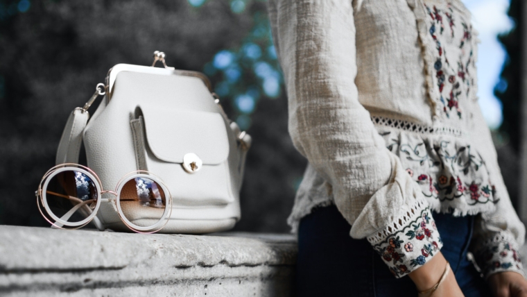 How to Build a Feminine Wardrobe on a Budget in 2023 1 woman wearing beige and red floral top leaning on gray concrete slab with white leather bag ontop