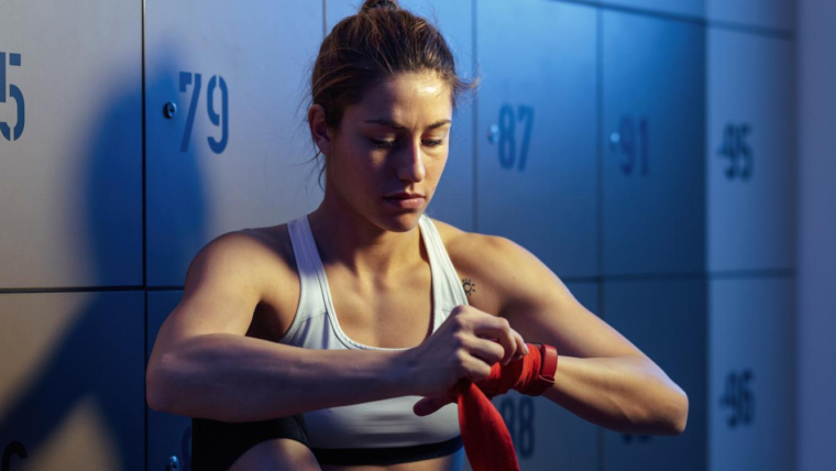 Les meilleures routines d'entraînement pour les femmes en 2024 1 jeune femme athlétique préparant un entraînement de boxe en enveloppant ses mains avec des sangles dans le vestiaire du gymnase